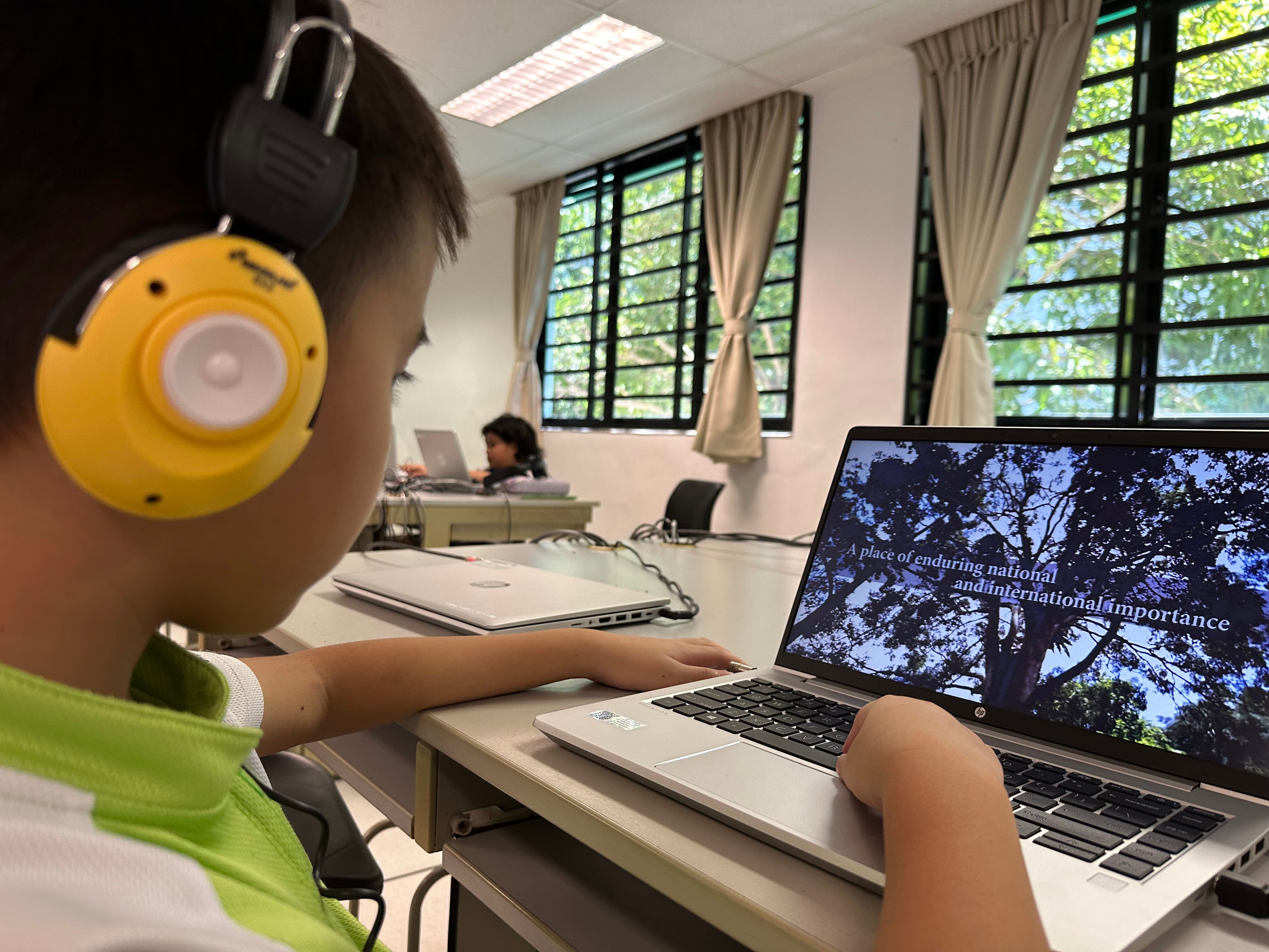 A child wearing yellow headphones watches a laptop showing trees with text about national and international importance.